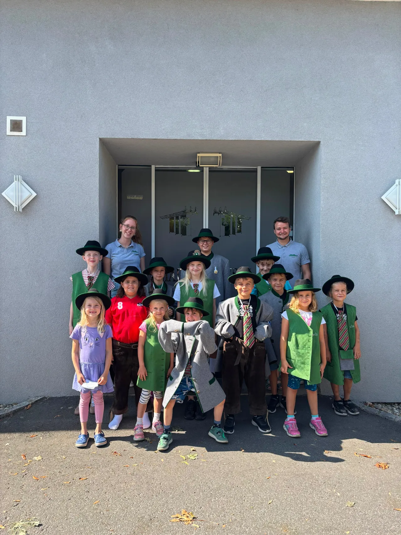 Eine Gruppe von Kindern und zwei Erwachsenen steht vor einem Gebäude, alle tragen grüne Westen und Hüte. Die Kinder tragen verschiedene Kleidungsstücke, und die Erwachsenen lächeln.