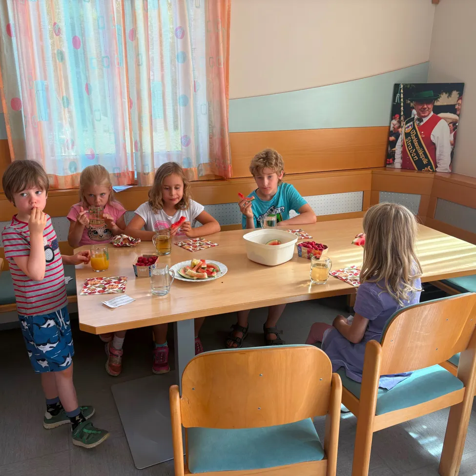 Sechs Kinder sitzen um einen Holztisch herum, essen Snacks und trinken Saft. Der Tisch ist mit Tellern, Schüsseln und Gläsern gedeckt. Ein Bilderrahmen hängt an der Wand hinter ihnen.