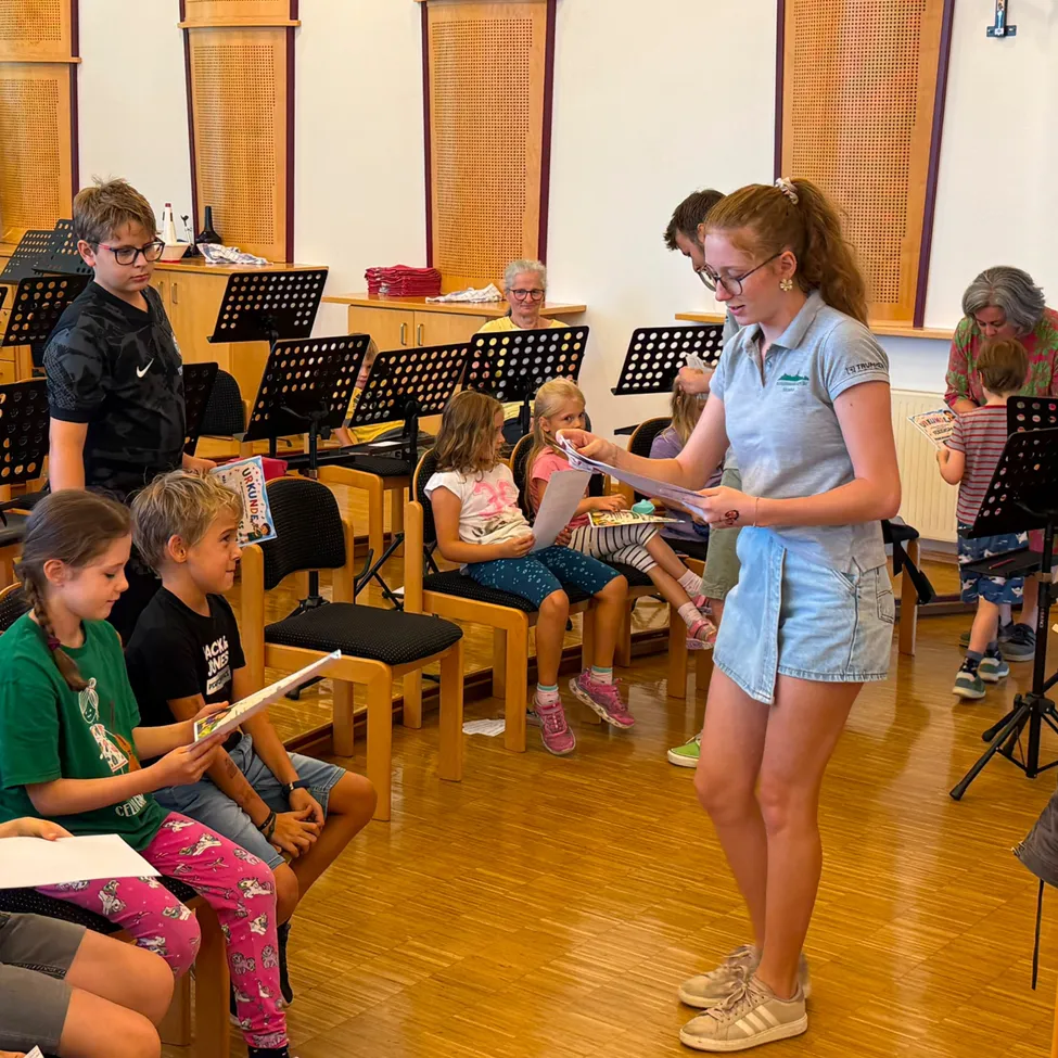 Eine junge Frau steht vor einer Gruppe von Kindern, hält ein Blatt Papier in der Hand und liest laut vor. Die Kinder sitzen auf Stühlen und hören aufmerksam zu, wobei im Hintergrund Musikinstrumente und Notenpapier zu sehen sind.
