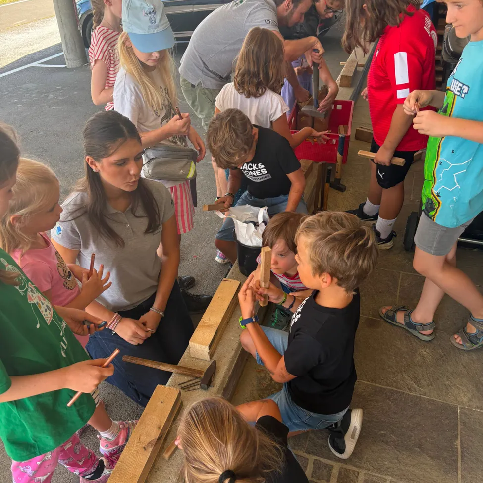 Eine Gruppe von Kindern und Erwachsenen versammelt sich um eine Holzstruktur, einige halten Hämmer und Meißel und nehmen möglicherweise an einer Bastelaktivität teil.