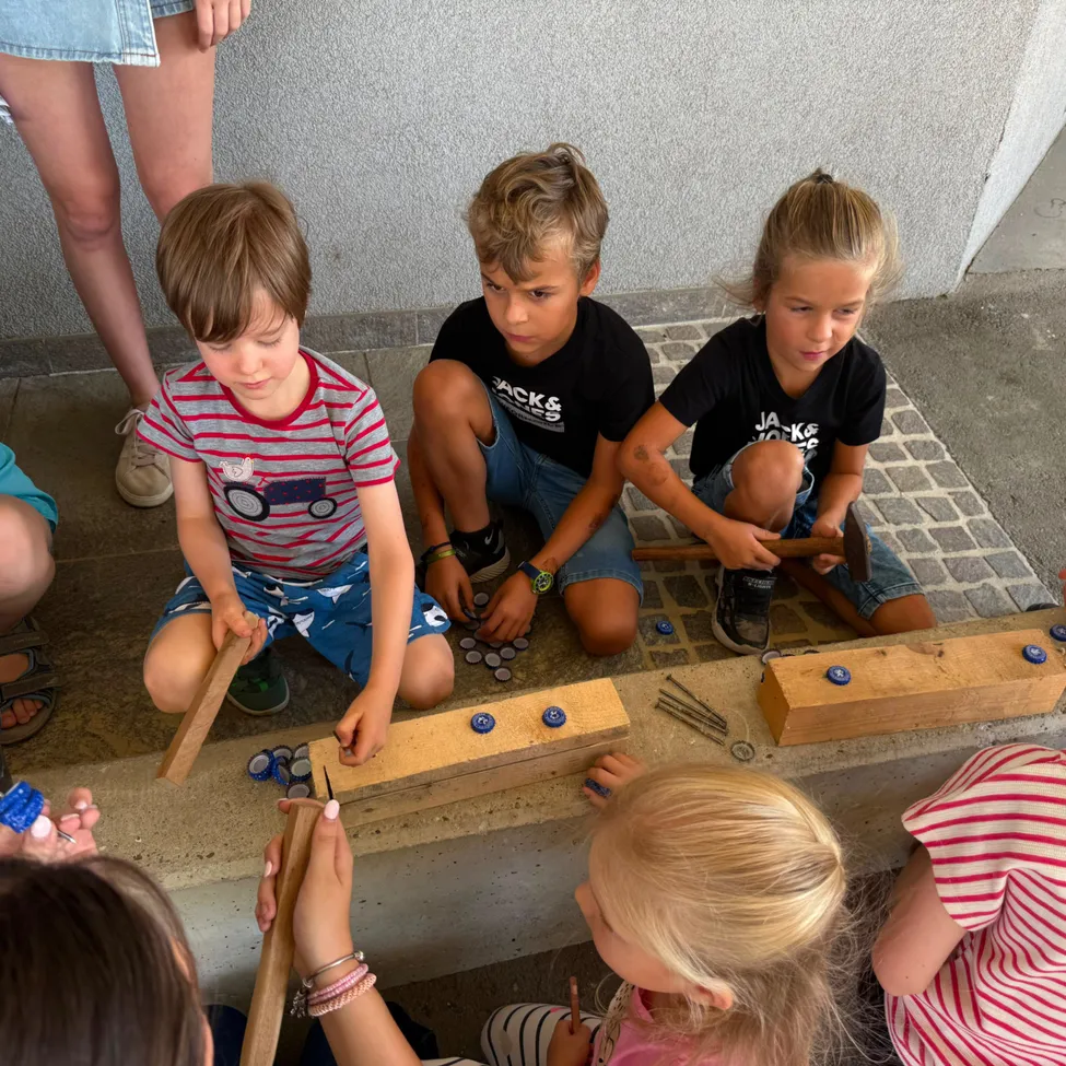 Eine Gruppe von Kindern sitzt auf dem Boden und ist in einer Aktivität vertieft. Sie halten Holzstäbe und scheinen Flaschenkappen zu hämmern. Die Kinder tragen Freizeitkleidung. Der Boden besteht aus Ziegelsteinen.