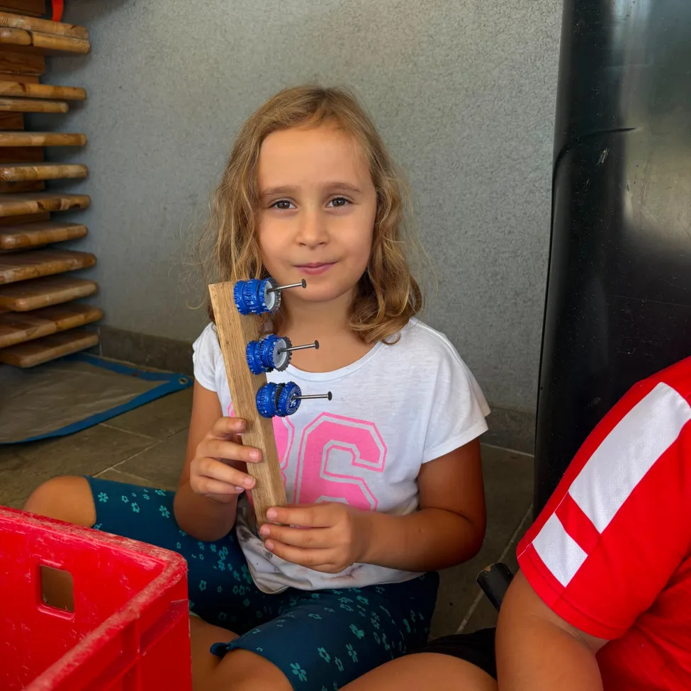 Ein junges Mädchen sitzt auf dem Boden, hält einen Holzstab mit blauen Zahnrädern und lächelt in die Kamera. Sie trägt ein weißes T-Shirt mit pinken Zahlen und blaue Shorts.