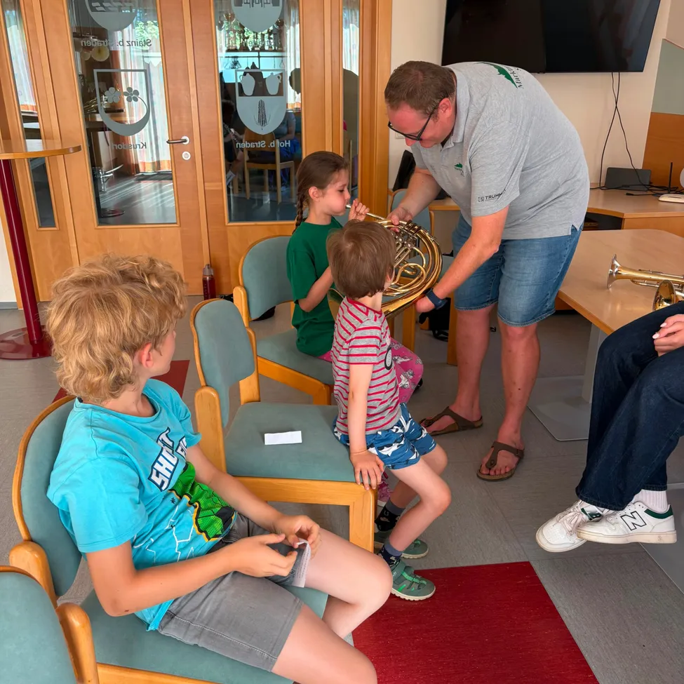 Ein Mann bringt Kindern das Spielen einer Posaune bei in einem Raum mit Holztüren und einem an der Wand montierten Fernseher.
