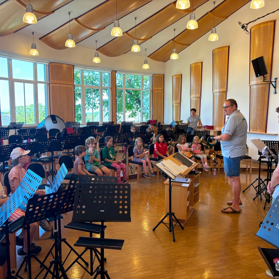 Ein Mann steht vor einer Gruppe von Kindern in einem Musikraum und hält einen Notenständer. Die Kinder sitzen auf Stühlen mit Notenständern vor ihnen.