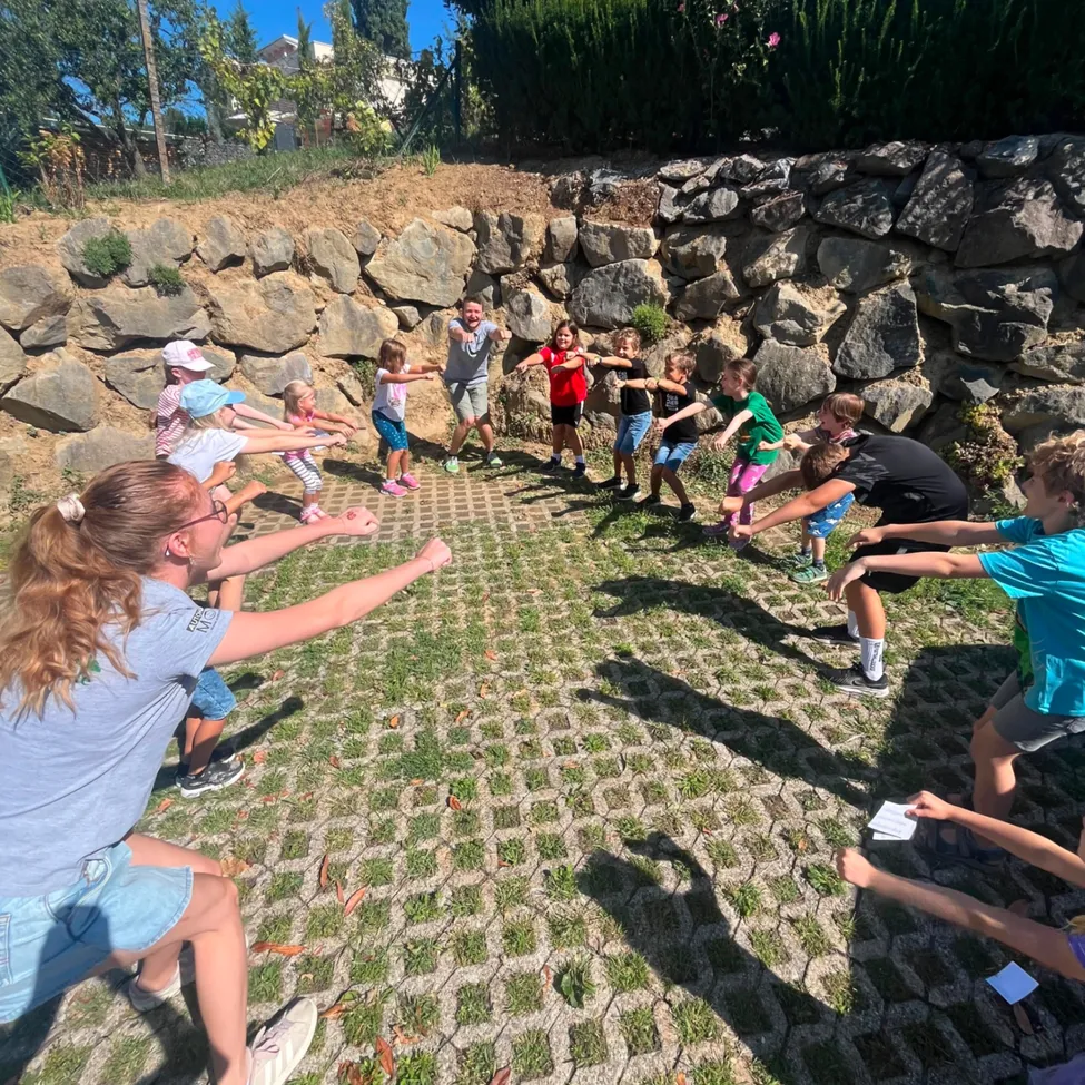 Eine Gruppe von Kindern und Erwachsenen spielt ein Spiel auf einem Rasenfeld mit einer Steinmauer im Hintergrund.