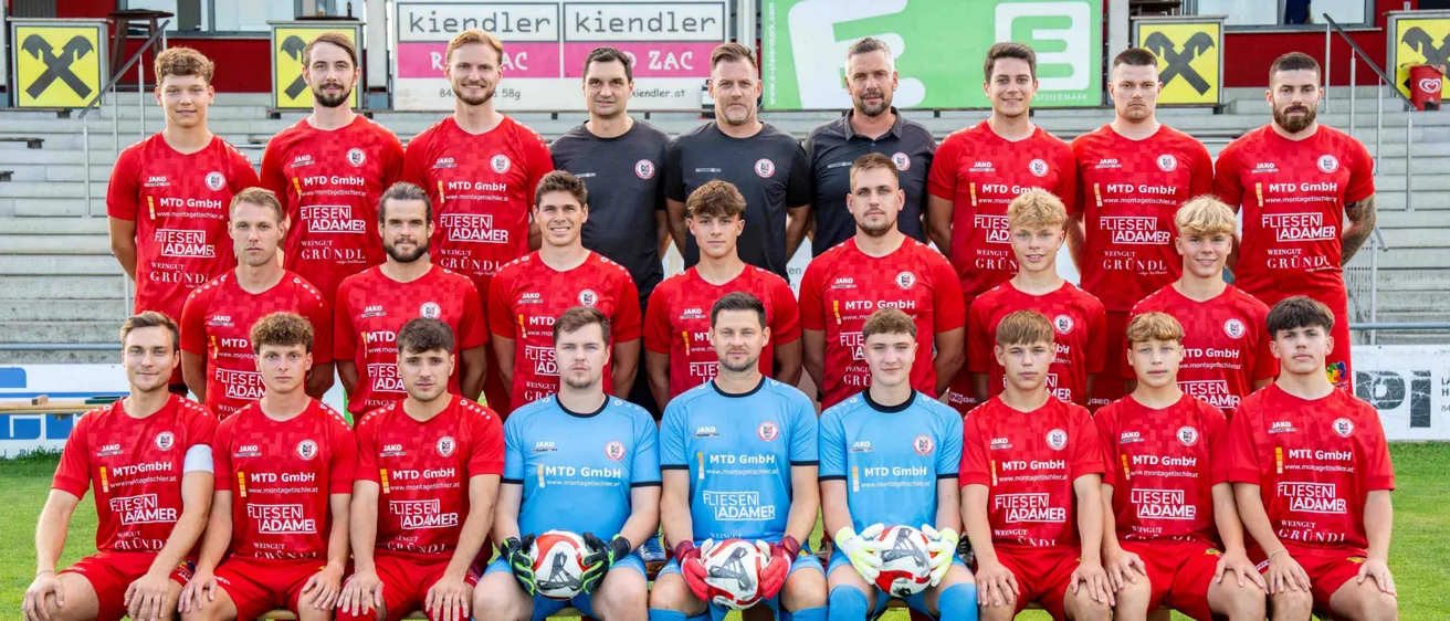 Eine Fußballmannschaft mit Spielern in roten und blauen Trikots posiert für ein Gruppenfoto auf dem Spielfeld. Im Hintergrund befinden sich Werbebanner mit verschiedenen Texten.