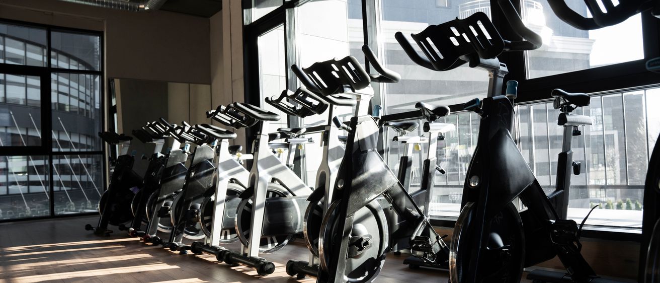 Eine Reihe von Trainingsrädern steht in einem Fitnessstudio mit großen Fenstern, die eine Stadtlandschaft zeigen. Die Fahrräder sind alle schwarz mit silbernen Rahmen, und Sonnenlicht scheint durch die Fenster.