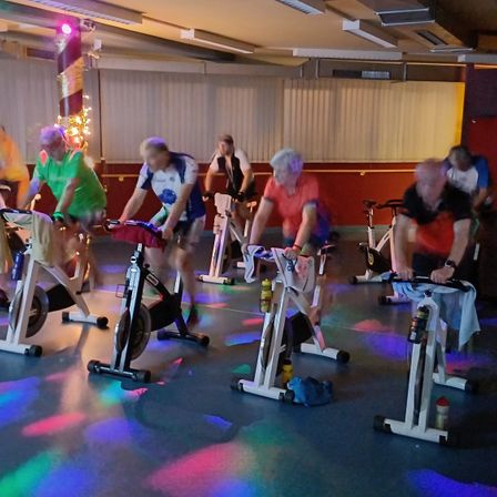 Eine Gruppe älterer Erwachsener trainiert auf Spinning-Bikes in einem Raum mit bunten Lichtern auf dem Boden.