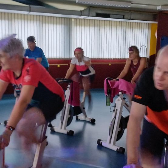 Mehrere Personen fahren in einem Fitnessstudio auf stationären Fahrrädern, mit weißen Wänden, Deckenleuchten und roten und gelben Wänden im Hintergrund.