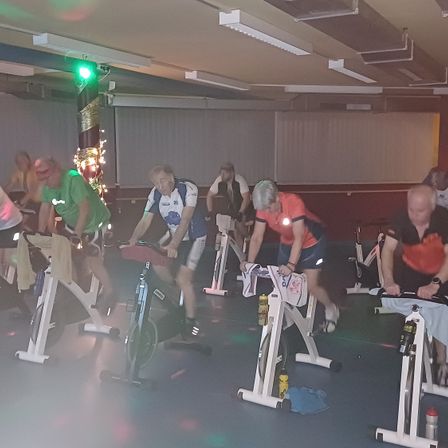 Eine Gruppe von Menschen trainiert auf Spinning-Bikes in einem Fitnessstudio. Sie tragen Sportbekleidung und einige haben reflektierende Ausrüstung. Das Fitnessstudio ist gut beleuchtet mit Deckenleuchten.