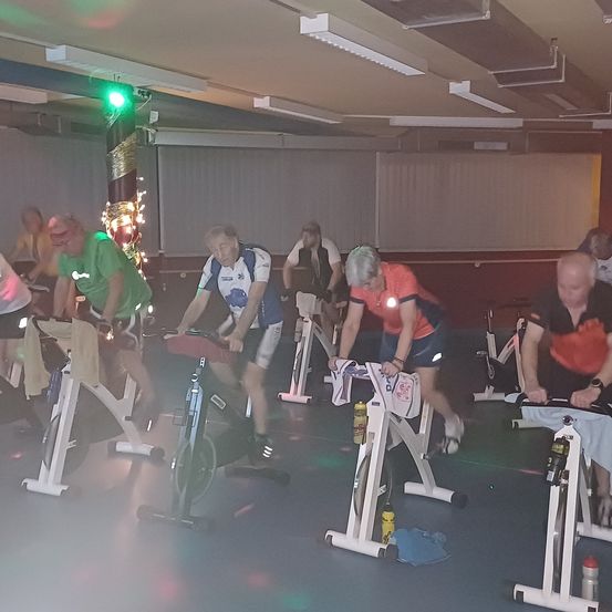 Eine Gruppe von Menschen trainiert auf Spinning-Bikes in einem Fitnessstudio. Sie tragen Sportbekleidung und einige haben reflektierende Ausrüstung. Das Fitnessstudio ist gut beleuchtet mit Deckenleuchten.