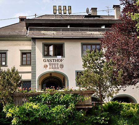 Ein großes weißes Gebäude namens Gasthof ist von Bäumen und Büschen umgeben. Es hat ein braunes Dach und Schornsteine. Es gibt eine Veranda mit Blumen und Pflanzen.