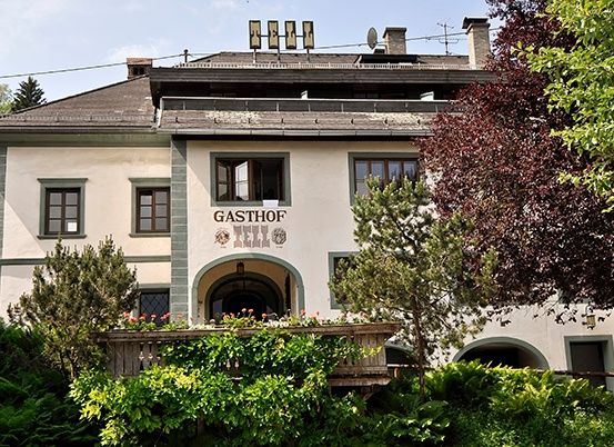 Ein großes weißes Gebäude namens Gasthof ist von Bäumen und Büschen umgeben. Es hat ein braunes Dach und Schornsteine. Es gibt eine Veranda mit Blumen und Pflanzen.