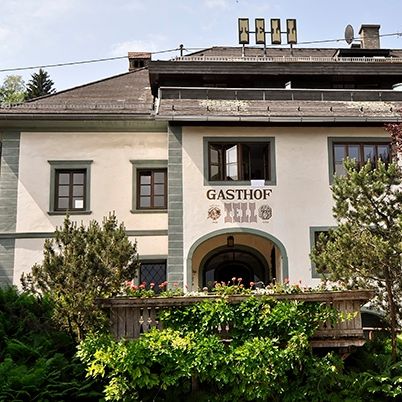 Ein weißes Gebäude mit der Aufschrift 'Gasthof' über dem Eingang ist von üppiger Grünfläche und Bäumen umgeben. Das Gebäude hat ein braunes Dach und mehrere Fenster.