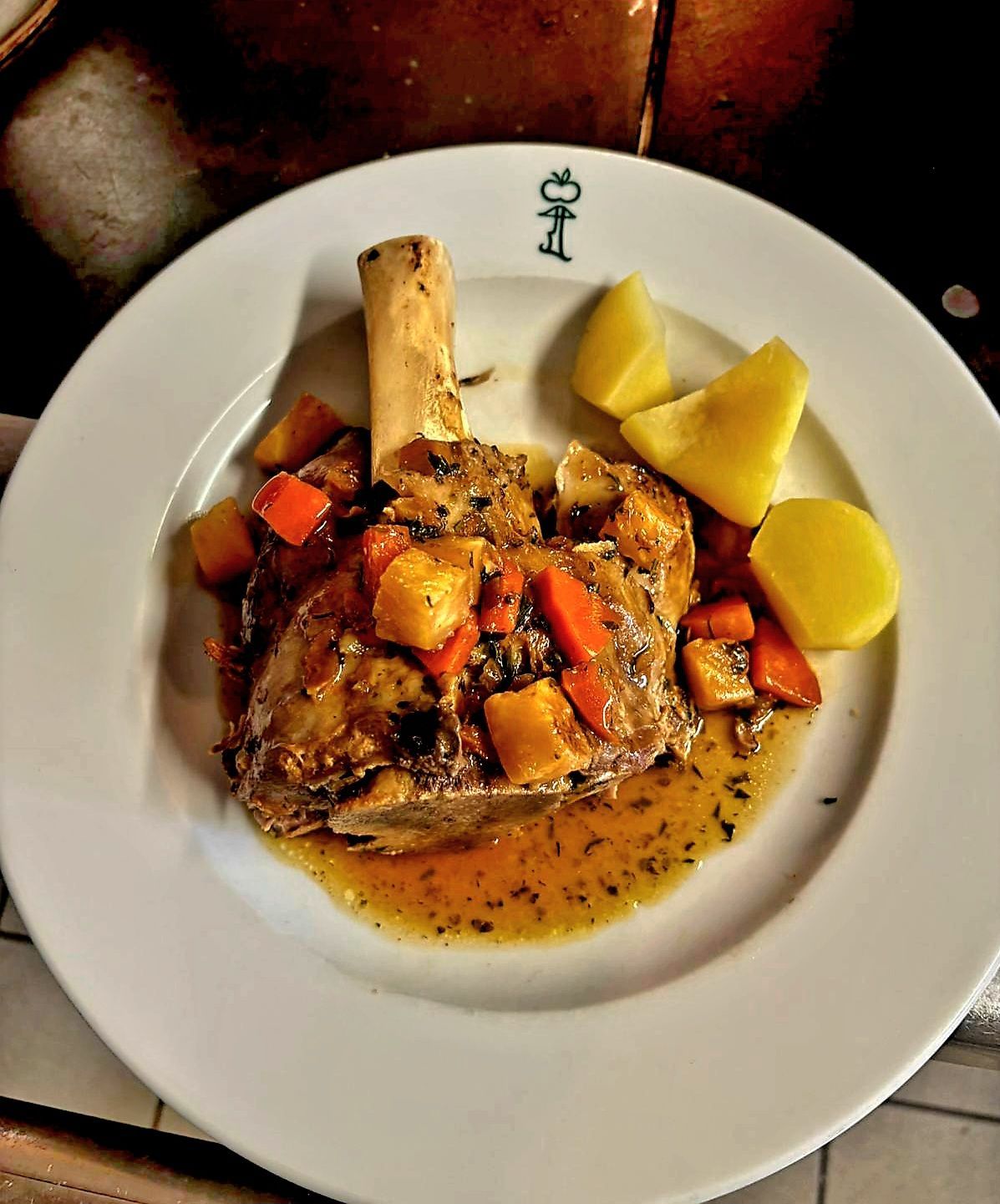 Ein Teller mit geschmorter Lammkeule, begleitet von gebratenen Kartoffeln und Karotten, auf einem weißen Teller mit einem Logo.