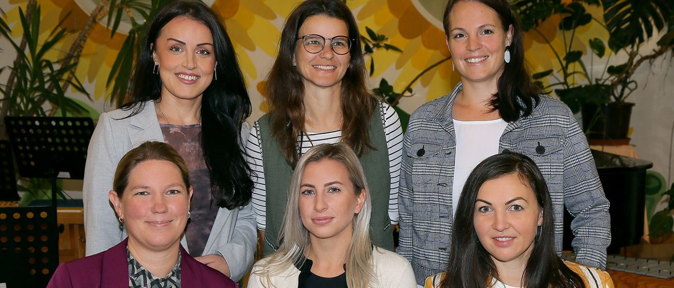 Sechs Frauen posieren für ein Gruppenfoto, lächelnd und vor einer gelben Blumenwand stehend.