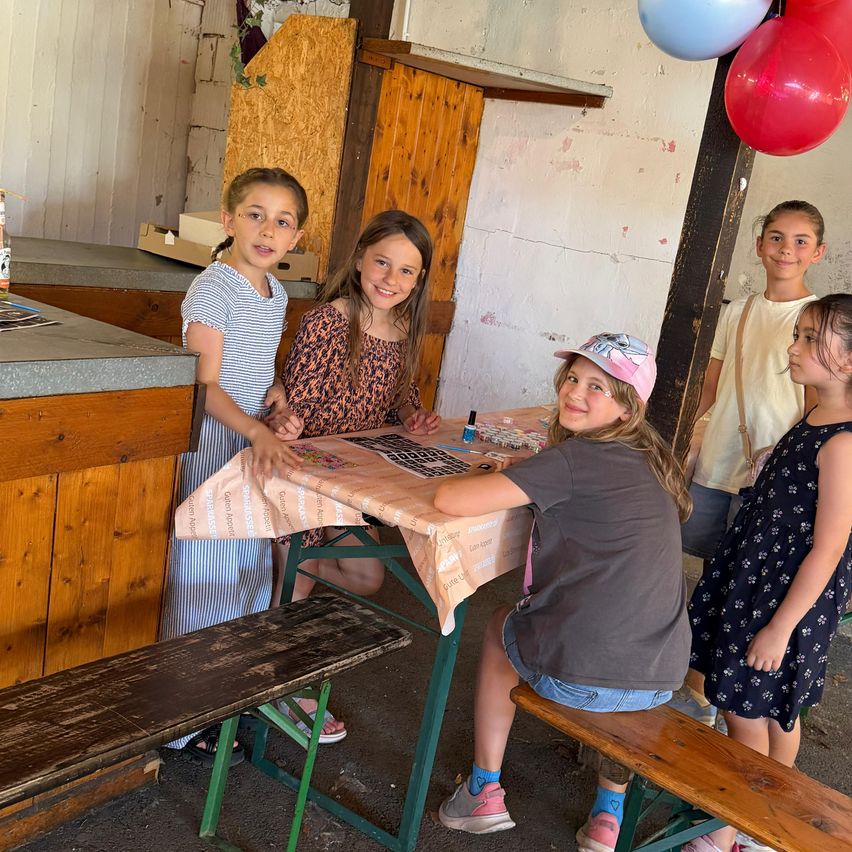 Fünf junge Mädchen stehen um einen Tisch herum, lächeln und schauen auf ein Spielbrett. Ein Mädchen sitzt auf einer Bank, während andere hinter dem Tisch stehen. Eine Theke mit einigen Gegenständen befindet sich links, und im Hintergrund sind Luftballons zu sehen.