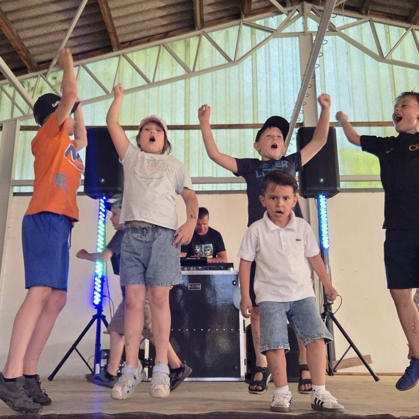 Mehrere Kinder tanzen und springen auf einer Bühne mit Musikgeräten. Einige tragen Hüte, und zwei haben ihre Hände erhoben. Ein DJ spielt Musik hinter ihnen.