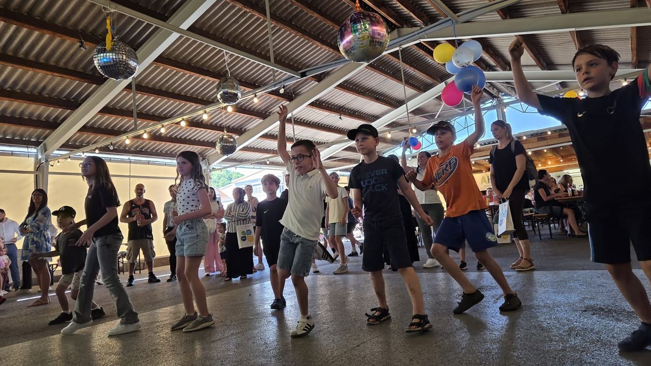 Eine Gruppe von Kindern tanzt gemeinsam in einem Saal mit Ballons und Discokugeln, die von der Decke hängen. Sie werden von anderen Menschen beobachtet, die sie umgeben.