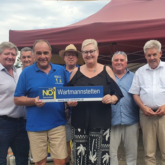 Eine Gruppe von Menschen steht unter einem Zelt und lächelt für ein Foto. Ein Mann hält ein blaues Schild mit der Aufschrift 'NO$ Wartmannstetten'.