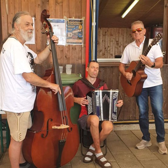 Drei Männer stehen in einem Raum und spielen jeweils ein Musikinstrument. Der Mann links hält ein Cello, der Mann in der Mitte spielt Akkordeon und der Mann rechts spielt Gitarre.