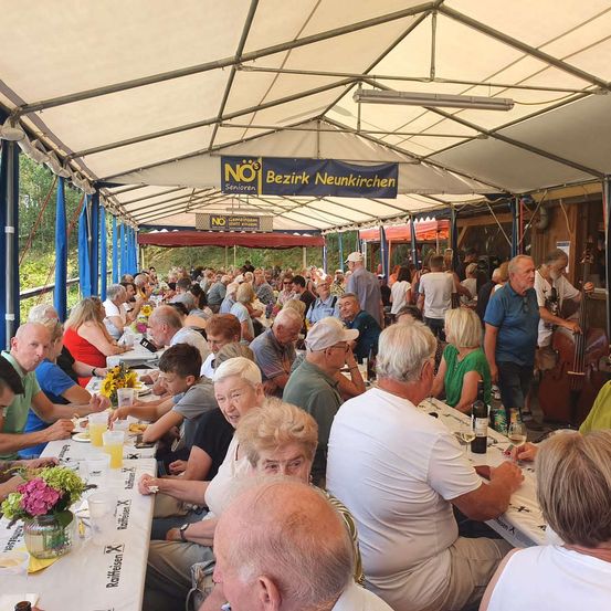 Eine Gruppe älterer Menschen diniert unter einem Zelt mit einem Banner, das 'NO Senioren Bezirk Neunkirchen' steht. Sie sitzen an langen Tischen mit Blumen und Weingläsern. Ein Cellist spielt im Hintergrund.