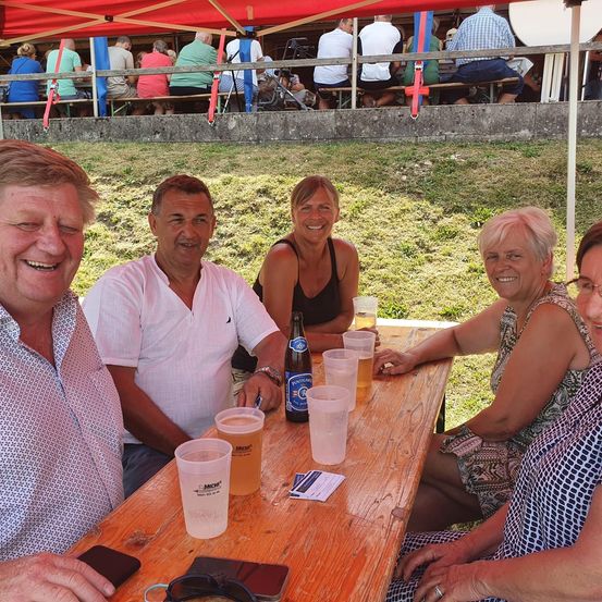Fünf Personen sitzen an einem Picknicktisch unter einem roten Baldachin, lächeln und posieren für ein Foto. Der Tisch ist mit Gläsern, einer Flasche und einer Karte bestückt. Hinter ihnen sitzen eine Gruppe von Menschen auf Bänken unter demselben Baldachin.