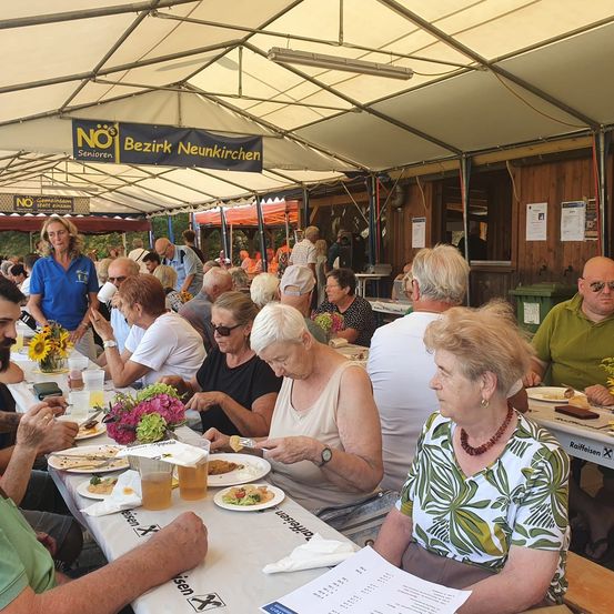 Eine Gruppe älterer Menschen sitzt an einem langen Tisch in einem Zelt, isst und trinkt. Ein Schild darüber steht 'NO Senioren'.