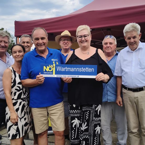 Eine Gruppe von Menschen steht unter einem Zelt und lächelt für ein Foto. Ein Mann in einem blauen Shirt hält ein blaues Schild mit dem Text 'NOJ Wartmannstettten'.