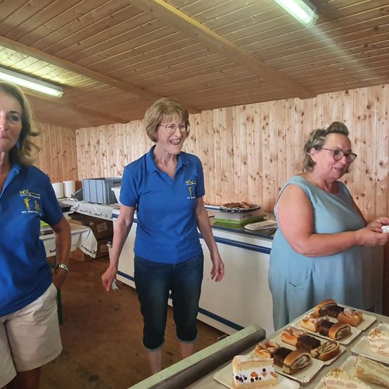 Drei Frauen in einem Raum mit Holzwänden, eine Frau in einem blauen Shirt lächelt und geht auf einen Tisch mit Gebäck zu.