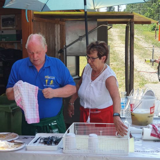 Ein älterer Mann und eine ältere Frau stehen hinter einem Tisch, der Mann hält ein Handtuch und die Frau einen Becher. Der Tisch ist mit Tellern, Bechern und einem Korb gedeckt. Die Umgebung scheint im Freien zu sein.