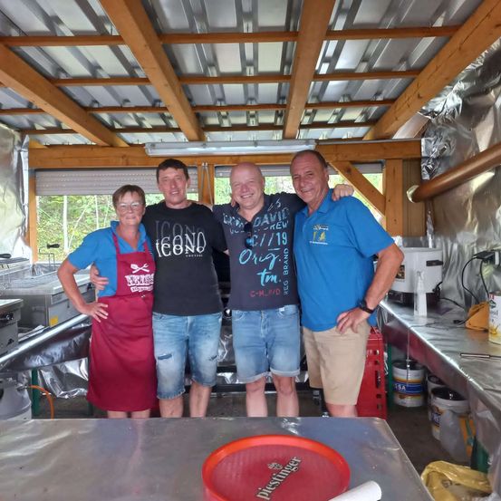Vier Personen stehen in einer Küche mit Metalldecke und Holzbalken. Sie lächeln und posieren für ein Foto. Die Frau trägt ein rotes Schürzen. Der Mann links trägt ein schwarzes T-Shirt mit Logo. Der Mann in der Mitte trägt ein blaues Hemd. Der Mann rechts trägt ein blaues Poloshirt. Auf dem Tisch steht ein roter Teller.