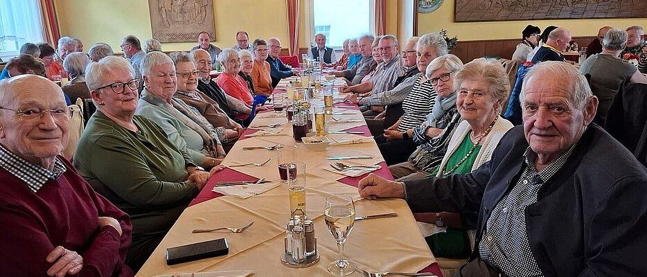 Eine Gruppe älterer Menschen sitzt an einem langen Tisch in einem Restaurant mit einem blauen Banner, das 'Wartmannstetten' im Vordergrund sagt.