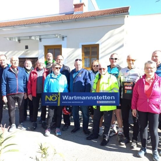 Eine Gruppe von Menschen in Radbekleidung posiert für ein Foto vor einem Haus mit einem Schild, das 'Wartmannstetten' steht.