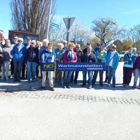 Eine Gruppe von Menschen in lässiger Outdoor-Kleidung steht auf einer Straße und hält ein Banner mit der Aufschrift 'Wartmannstetten'. Sie lächeln und scheinen für ein Foto zu posieren.