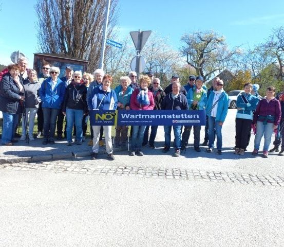 Eine Gruppe von Menschen in lässiger Outdoor-Kleidung steht auf einer Straße und hält ein Banner mit der Aufschrift 'Wartmannstetten'. Sie lächeln und scheinen für ein Foto zu posieren.