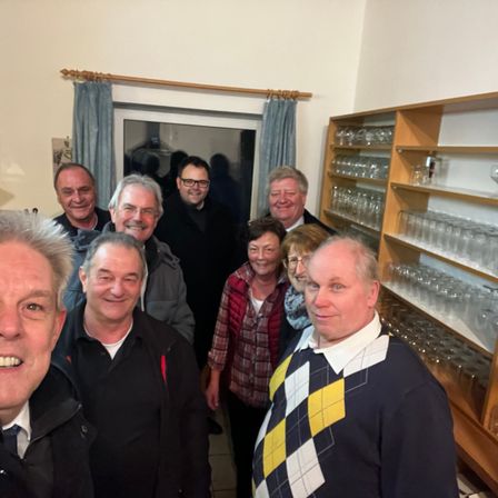Eine Gruppe von Menschen, darunter Männer und Frauen, steht in einem Raum und lächelt für ein Foto. Hinter ihnen befinden sich Regale mit Gläsern und ein Fenster mit Vorhängen.