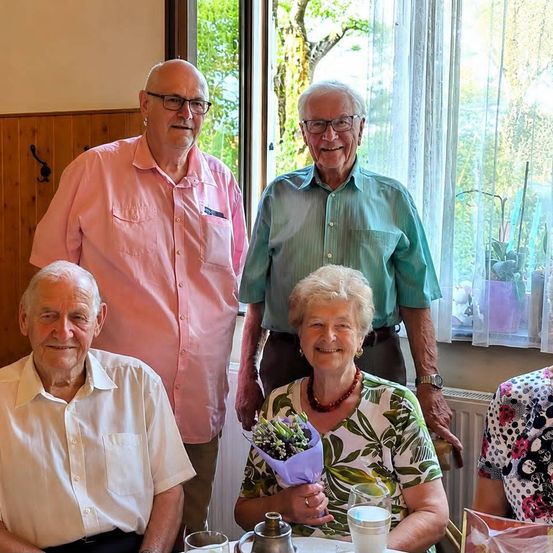 Vier ältere Personen, zwei Männer und zwei Frauen, lächeln und posieren für ein Foto in einem Raum mit Fenster und Heizkörper.
