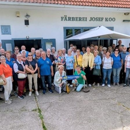 Eine Gruppe von Menschen, wahrscheinlich Touristen, posiert für ein Foto vor einem Gebäude mit dem Namen 'Färberei Josef Koo'. Sie sind lässig gekleidet und einige tragen Taschen. Das Gebäude hat ein Ziegeldach und Glasfenster.