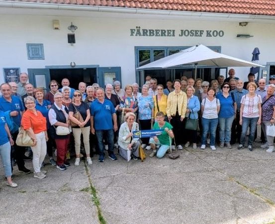 Eine Gruppe von Menschen, wahrscheinlich Touristen, posiert für ein Foto vor einem Gebäude mit dem Namen 'Färberei Josef Koo'. Sie sind lässig gekleidet und einige tragen Taschen. Das Gebäude hat ein Ziegeldach und Glasfenster.