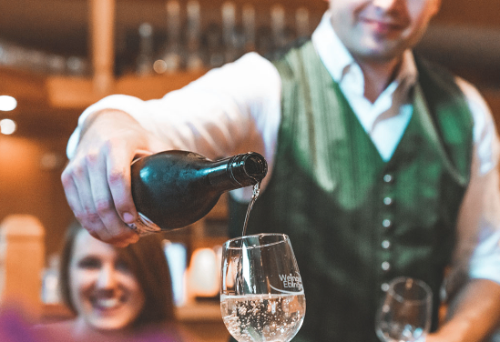 Ein Barkeeper gießt Wein aus einer Flasche in ein Weinglas, während eine lächelnde Frau zuschaut. Der Schauplatz ist drinnen, mit hölzernen Regalen im Hintergrund.