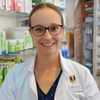 Eine Frau in einem weißen Laborkittel steht in einer Apotheke und lächelt. Hinter ihr sind Regale mit verschiedenen Medikamenten und Produkten. Sie trägt eine Brille und einen Ausweis, der 'Apotheker' steht.