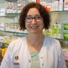 Eine Frau mit Brille und weißem Laborkittel steht in einer Apotheke. Hinter ihr stehen Regale mit verschiedenen Medikamenten, darunter Antistax, Sinupret und Cete.