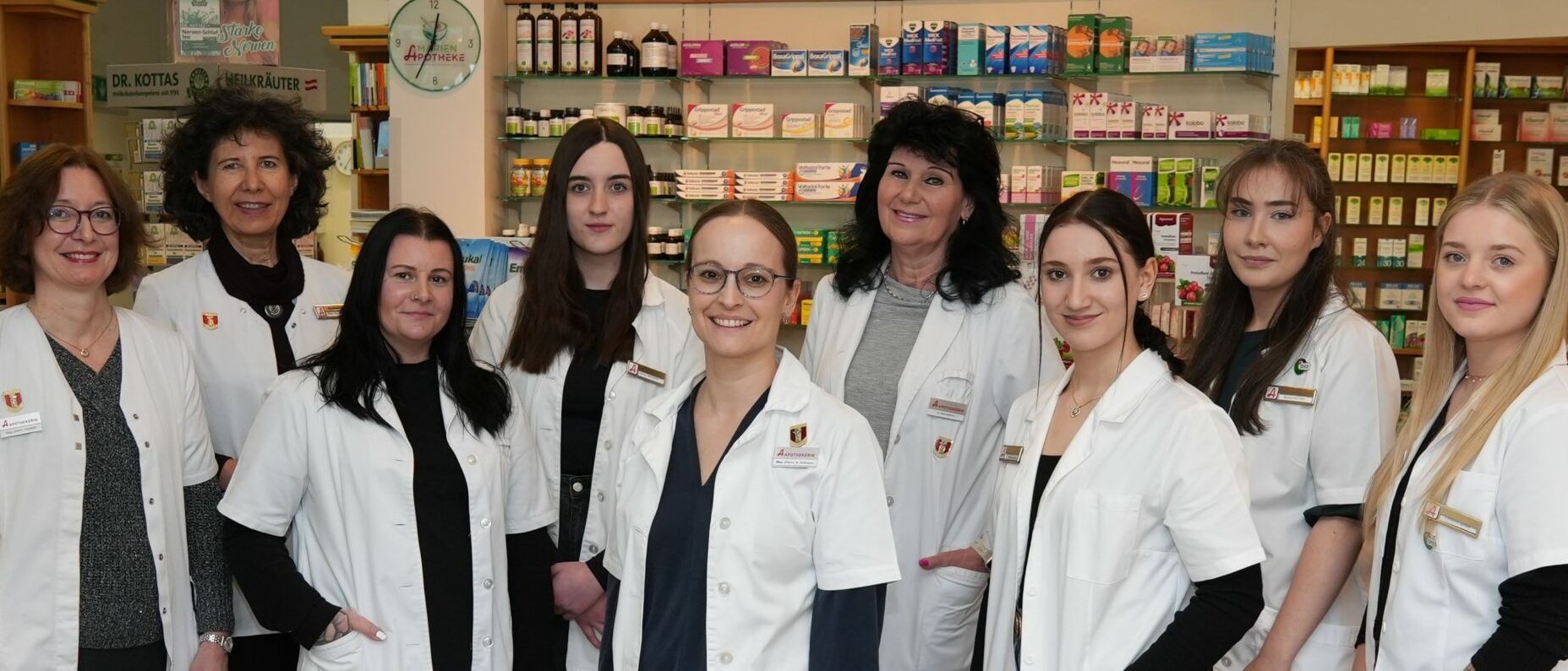 Fünf Frauen in weißen Laborkitteln stehen in einer Apotheke. Die Regale hinter ihnen sind mit verschiedenen Medikamenten bestückt. Ein Schild über den Regalen steht Schmerz Frei und Erkältung.