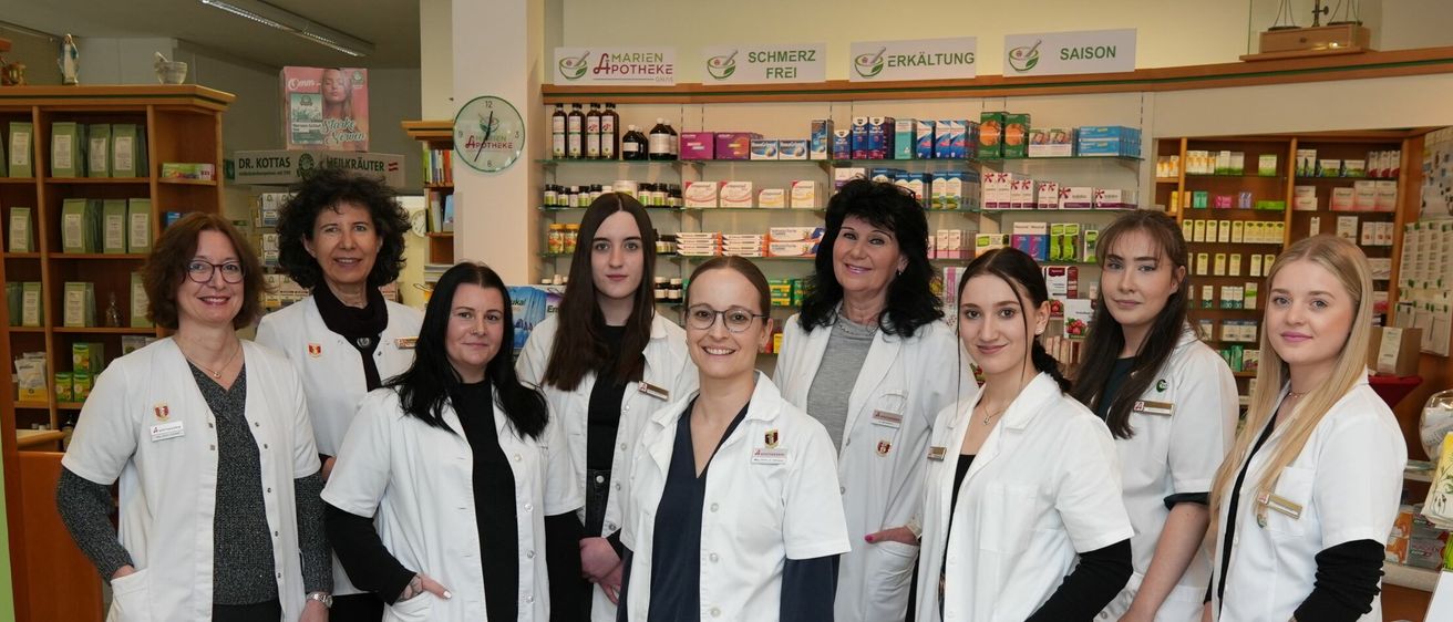 Fünf Frauen in weißen Laborkitteln stehen in einer Apotheke. Die Regale hinter ihnen sind mit verschiedenen Medikamenten bestückt. Ein Schild über den Regalen steht Schmerz Frei und Erkältung.