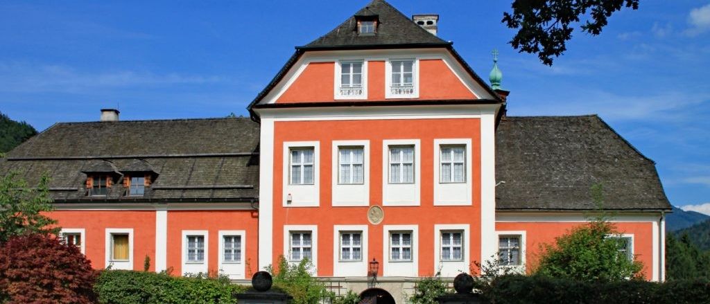 Ein großes, zweistöckiges Gebäude mit einem grauen Dach und roten Wänden. Der Eingang ist gewölbt, und es gibt Fenster im oberen Stockwerk. Das Gebäude ist von grünen Sträuchern umgeben.