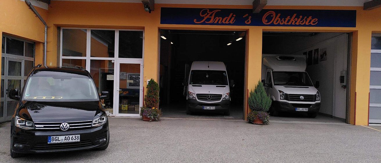 Ein schwarzer Wagen ist vor einem Gebäude namens 'Andi's Obstkiste' geparkt. Zwei weiße Lieferwagen sind im Garage geparkt.