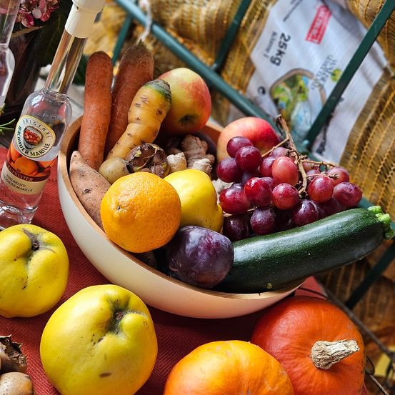 Eine Schüssel mit gemischtem Obst und Gemüse, darunter Äpfel, Orangen und Trauben. Es gibt auch eine Flasche Likör und einen Strauß Kräuter auf dem Tisch.
