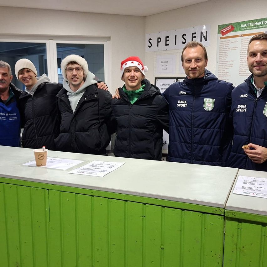 Fünf Männer stehen hinter einer Theke und lächeln für die Kamera. Sie tragen Wintermäntel und einige haben Weihnachtsmützen. Ein Becher und Papiere sind auf der Theke.