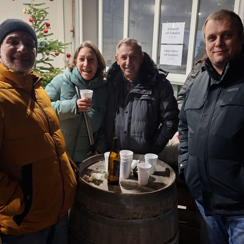 Vier Menschen posieren für ein Foto vor einem Fass mit Tassen, einer Flasche und einem Weihnachtsbaum. Der Mann rechts trägt eine Mütze und der Mann links einen Mantel.
